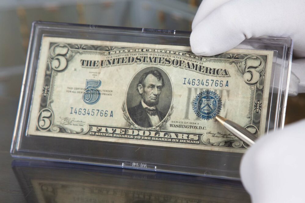 $5 US Treasury Blue Seal Silver Certificate in protective case, White-gloved hand holding it with pointer showing Blue Seal.